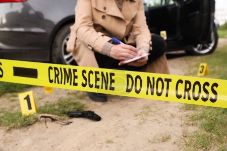 Professional detective examining crime scene outdoors, focus on yellow tape