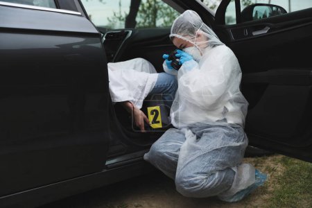 Photo for Criminologist taking photo of evidence at crime scene with dead body in car - Royalty Free Image