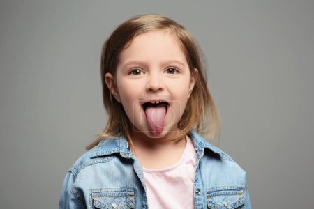 Téléchargez les photos : Petite fille émotionnelle montrant sa langue sur fond gris - en image libre de droit