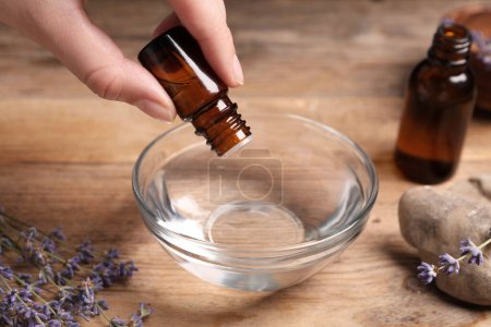 Photo for Woman dripping lavender essential oil from bottle into bowl at wooden table, closeup - Royalty Free Image