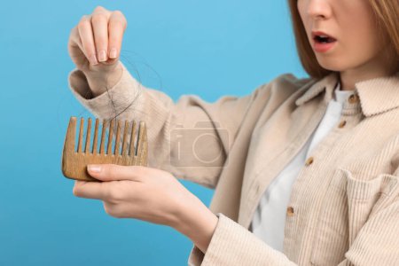 Woman untangling her lost hair from comb on light blue background, closeup. Alopecia problem
