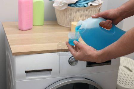 Homme versant adoucisseur de tissu de la bouteille dans le bouchon près de la machine à laver à l'intérieur, gros plan