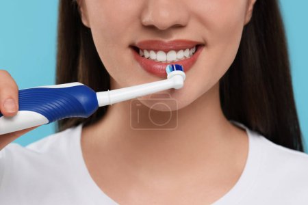 Woman brushing her teeth with electric toothbrush on light blue background, closeup