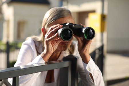 Téléchargez les photos : Concept de vie privée. Femme âgée curieuse avec des jumelles espionnant les voisins au-dessus de la clôture à l'extérieur - en image libre de droit