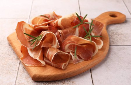 Slices of tasty cured ham and rosemary on tiled table, closeup