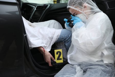 Photo for Criminologist taking photo of evidence at crime scene with dead body in car - Royalty Free Image