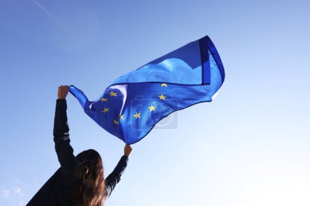 Mujer sosteniendo bandera de la Unión Europea contra el cielo azul al aire libre, vista de ángulo bajo