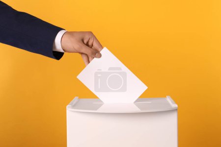 Photo for Man putting his vote into ballot box on yellow background, closeup - Royalty Free Image