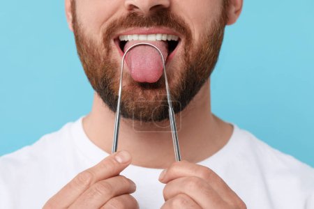 Man brushing his tongue with cleaner on light blue background, closeup