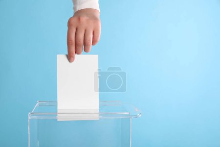 Woman putting her vote into ballot box on light blue background, closeup. Space for text