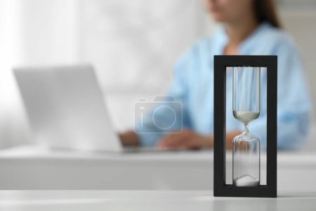 Hourglass with flowing sand on white table. Woman using laptop indoors, selective focus