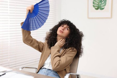 Foto de Joven mujer de negocios agitando ventilador de mano azul para refrescarse en la mesa en la oficina - Imagen libre de derechos