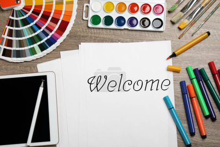 Téléchargez les photos : Papier avec mot Bienvenue, marqueurs colorés, aquarelle, palette et tablette sur table en bois, pose plate - en image libre de droit