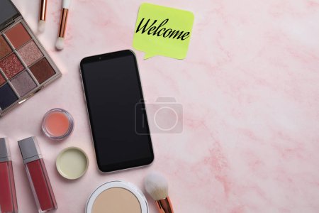 Téléchargez les photos : Note collante avec mot Bienvenue près du smartphone et des produits de maquillage sur table rose, pose plate - en image libre de droit