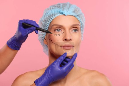 Frau bereitet sich auf Schönheitschirurgie vor, rosa Hintergrund. Arzt zeichnet Zeichnungen auf ihr Gesicht, Nahaufnahme