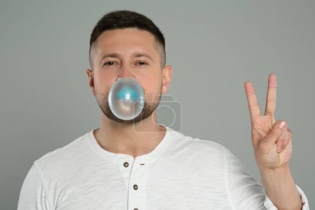 Téléchargez les photos : Bel homme soufflant chewing-gum bulle et montrant geste de paix sur fond gris clair - en image libre de droit