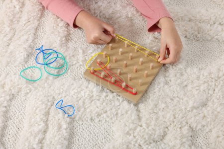 Motor skills development. Girl playing with geoboard and rubber bands on carpet, closeup