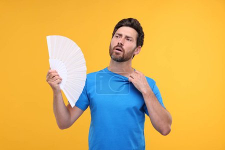 Foto de Hombre infeliz con ventilador de mano sufriendo de calor sobre fondo naranja - Imagen libre de derechos