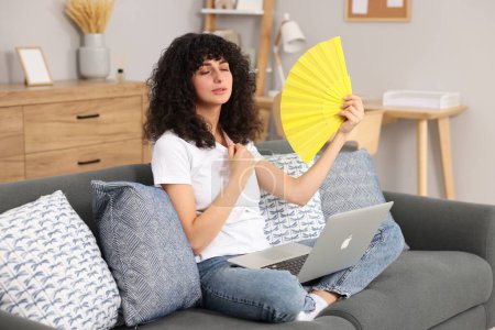 Foto de Mujer joven con portátil agitando ventilador de mano amarillo para refrescarse en el sofá en casa - Imagen libre de derechos
