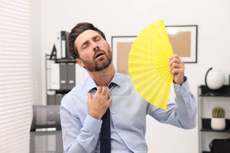 Foto de Barbudo hombre de negocios saludando ventilador de mano amarilla para refrescarse en la oficina - Imagen libre de derechos