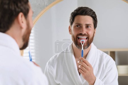 Handsome man brushing his tongue with cleaner near mirror in bathroom
