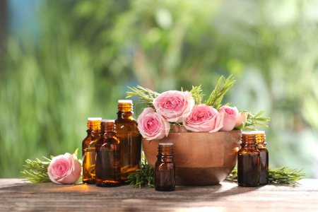 Foto de Botellas con aceites esenciales, rosas y romero sobre mesa de madera sobre fondo verde borroso. Espacio para texto - Imagen libre de derechos
