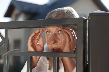 Concept of private life. Curious senior woman spying on neighbours over fence outdoors