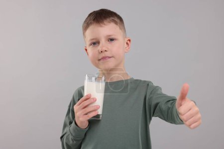 Téléchargez les photos : Mignon garçon avec verre de lait frais montrant pouce vers le haut sur fond gris clair - en image libre de droit