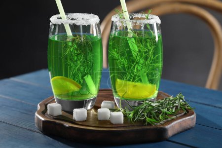 Photo for Glasses of homemade refreshing tarragon drink, sprigs and sugar cubes on blue wooden table, closeup - Royalty Free Image