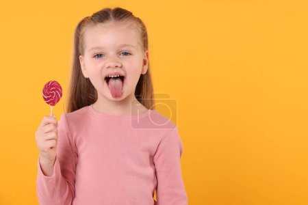 Funny little girl with bright lollipop swirl showing tongue on orange background, space for text