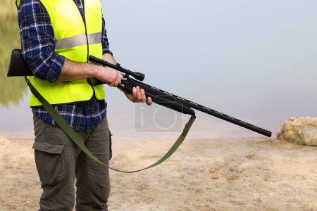 Photo for Man with hunting rifle wearing safety vest near lake outdoors, closeup - Royalty Free Image