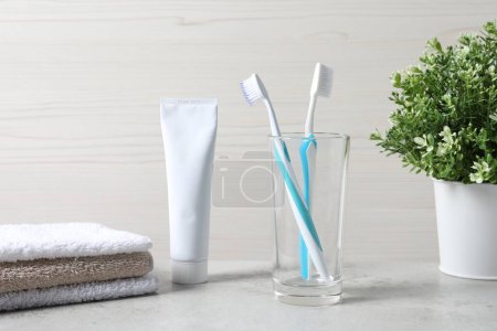 Téléchargez les photos : Brosses à dents en plastique dans un support en verre, tube de dentifrice et serviettes sur table gris clair - en image libre de droit
