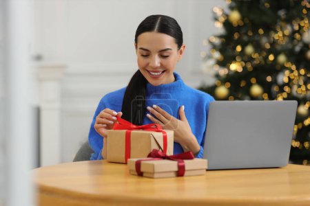 Foto de Celebrando la Navidad en línea con regalos intercambiados por correo. Mujer sonriente abriendo caja de regalo durante la videollamada en el ordenador portátil en casa - Imagen libre de derechos