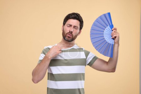 Foto de Hombre infeliz con ventilador de mano sufriendo de calor sobre fondo beige - Imagen libre de derechos