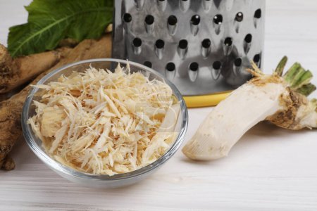 Photo for Grated horseradish in bowl, roots and grater on white wooden table, closeup - Royalty Free Image