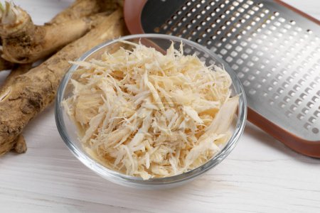 Photo for Grated horseradish in bowl, roots and hand grater on white wooden table, closeup - Royalty Free Image