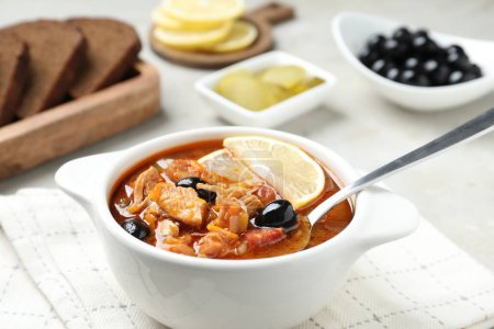 Meat solyanka soup with sausages, olives, vegetables and spoon in bowl on white table, closeup