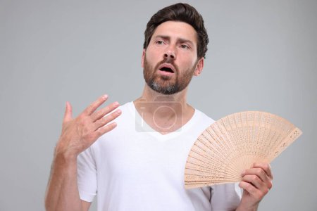 Foto de Hombre infeliz con ventilador de mano sufriendo de calor sobre fondo gris claro - Imagen libre de derechos