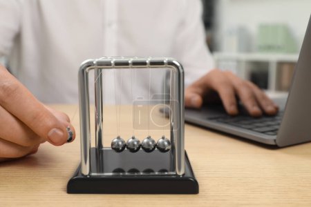 Photo for Man playing with Newton's cradle and using laptop at wooden table, closeup. Physics law of energy conservation - Royalty Free Image
