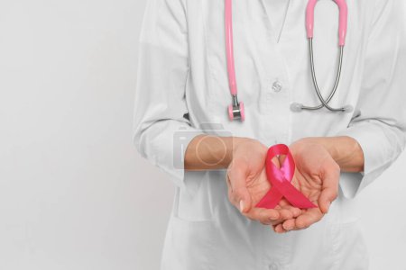 Téléchargez les photos : Médecin avec stéthoscope tenant un ruban rose sur fond blanc, gros plan et espace pour le texte. Sensibilisation au cancer du sein - en image libre de droit