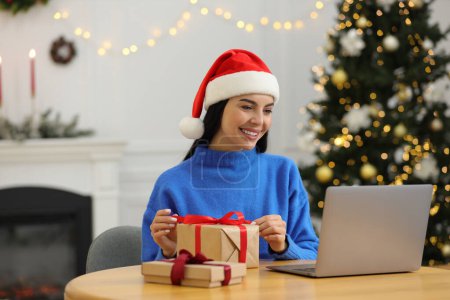 Foto de Celebrando la Navidad en línea con regalos intercambiados por correo. Mujer sonriente en la caja de regalo de apertura del sombrero de Santa durante la videollamada en el ordenador portátil en casa - Imagen libre de derechos