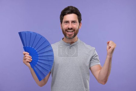 Foto de Hombre emocional sosteniendo ventilador de mano sobre fondo púrpura - Imagen libre de derechos