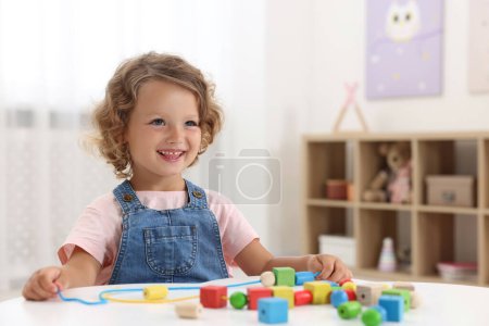 Motorische Entwicklung. Kleines Mädchen spielt mit Holzstücken und Schnur zum Einfädeln am Tisch im Haus