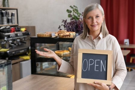 Photo for Smiling business owner holding open sign and inviting to come into her cafe, space for text - Royalty Free Image