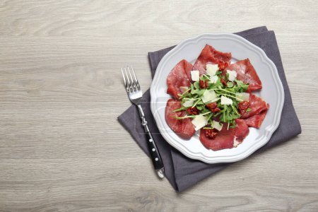 Photo for Plate of tasty bresaola salad with sun-dried tomatoes, parmesan cheese and fork on wooden table, top view. Space for text - Royalty Free Image