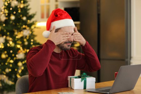 Celebrando la Navidad en línea con regalos intercambiados por correo. Hombre en Santa sombrero cubriendo los ojos antes de abrir la caja de regalo durante la videollamada en el ordenador portátil en casa