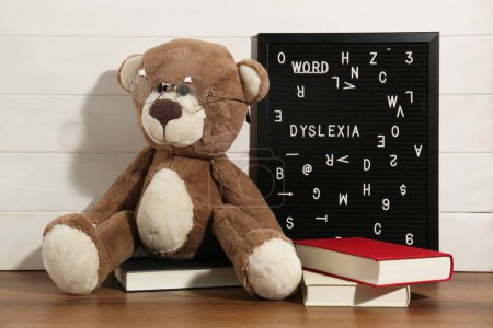 Tablero de letras negro con palabra Dislexia, libros y osito de peluche en mesa de madera cerca de la pared blanca