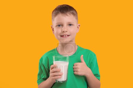 Téléchargez les photos : Mignon garçon avec un verre de lait frais montrant pouce vers le haut sur fond orange - en image libre de droit