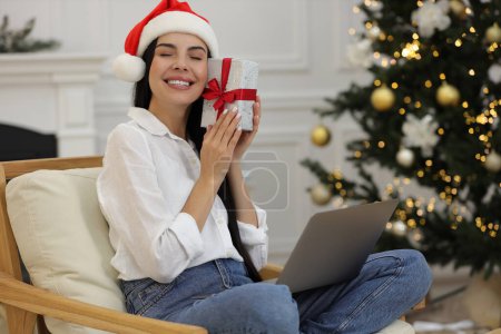 Celebrando la Navidad en línea con regalos intercambiados por correo. Mujer sonriente en sombrero de Santa con caja de regalo durante la videollamada en el ordenador portátil en casa