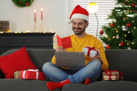 Celebrating Christmas online with exchanged by mail presents. Man in Santa hat with greeting card and gift box during video call on laptop at home